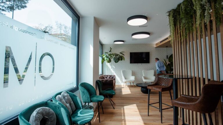 Sala de espera de OM Clinics, clínica dental en Alcorcón, con butacas verdes junto al ventanal, listones de madera y plantas colgantes
